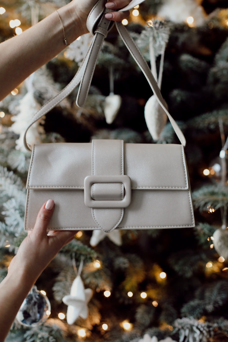 Close-up of a beige leather handbag with a statement buckle and adjustable strap, held in front of a decorated Christmas tree with warm lights and ornaments, perfect for festive or elegant occasions.