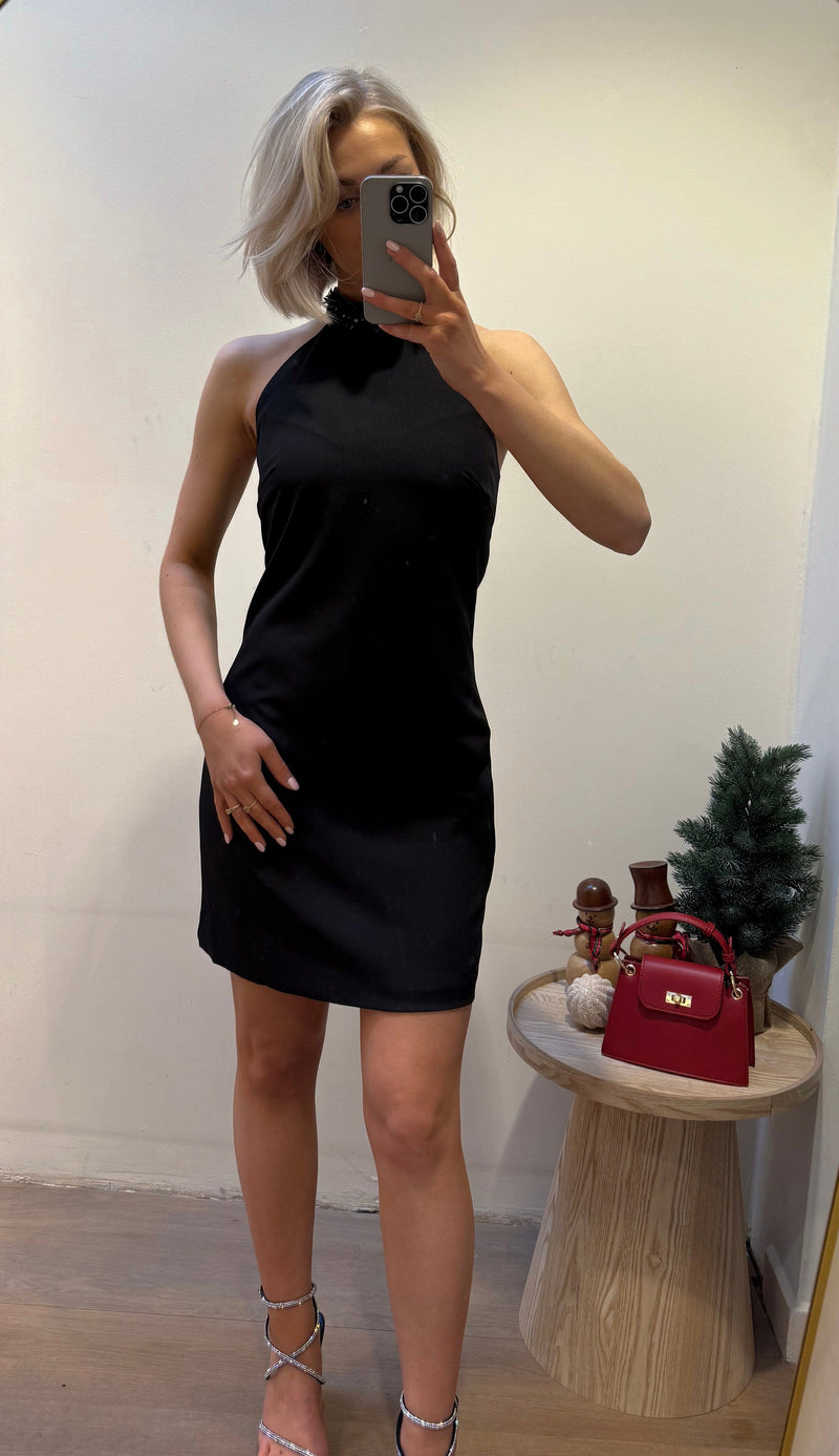 Model showcasing a sleek black halter-neck dress with a fitted silhouette, perfect for elegant evening events or formal occasions, styled with subtle accessories and a festive background